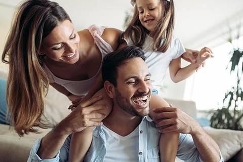 Family of three playing together at home, with father holding daughter on shoulders while mother hugs them from behind
